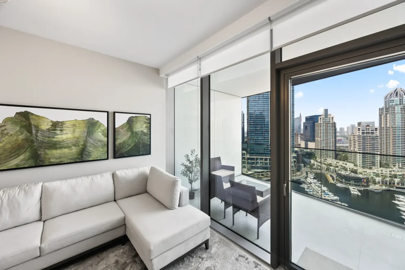 Modern living room with a white sectional sofa, framed landscape art, and a glass balcony door overlooking a marina and city skyline.