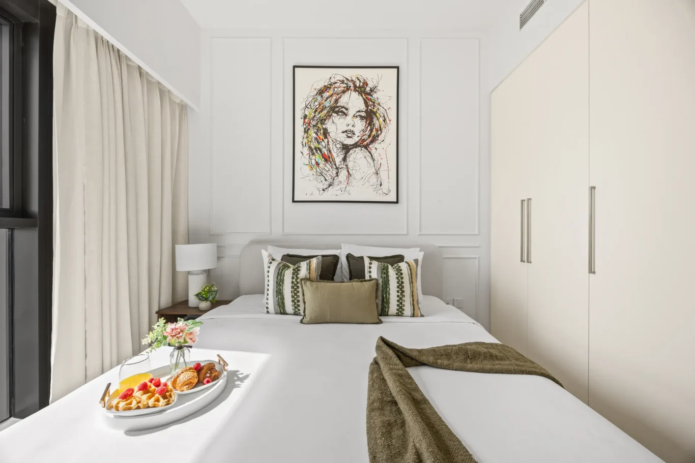 Hotel bedroom with white walls, large bed, and framed abstract portrait above the headboard; breakfast tray with pastries on the bed.