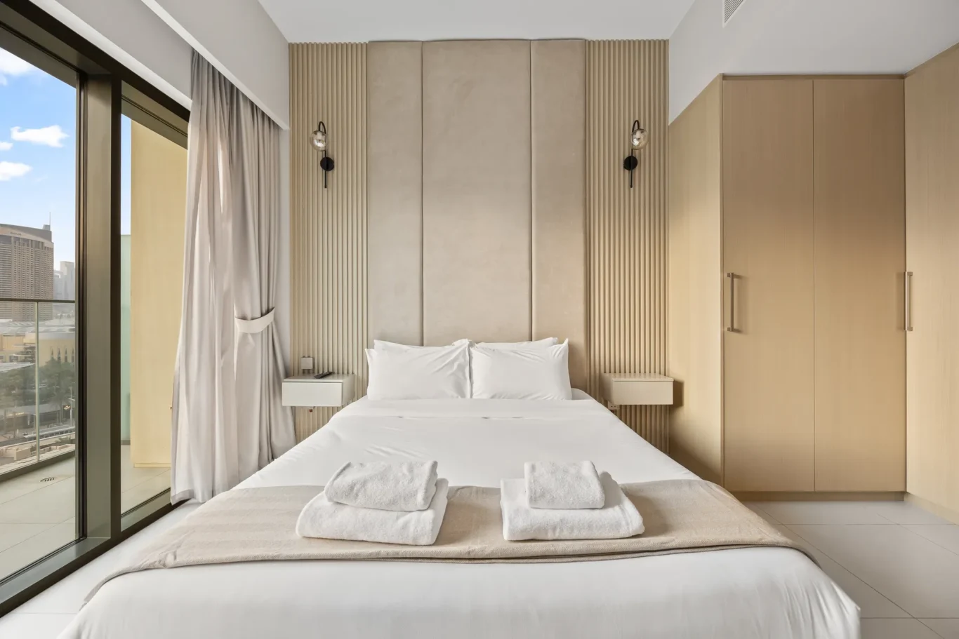 Elegant hotel bedroom with a padded beige headboard wall, wall lamps, and a large glass balcony window overlooking the city; bed with white linens.