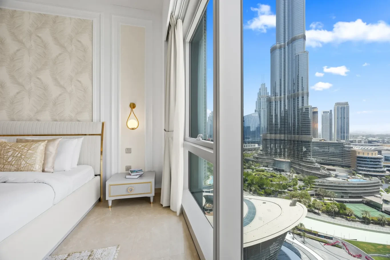 Luxurious hotel bedroom with white-gold decor and a large window overlooking a modern city skyline of glass skyscrapers.",