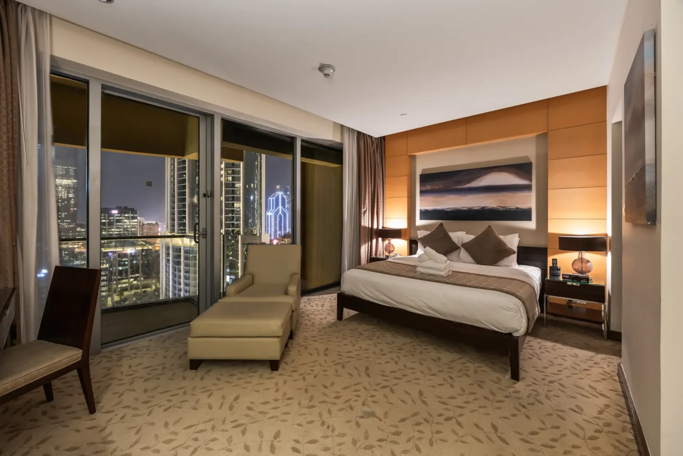 Hotel room at night with a king bed, seating area, and city skyline visible through large windows.