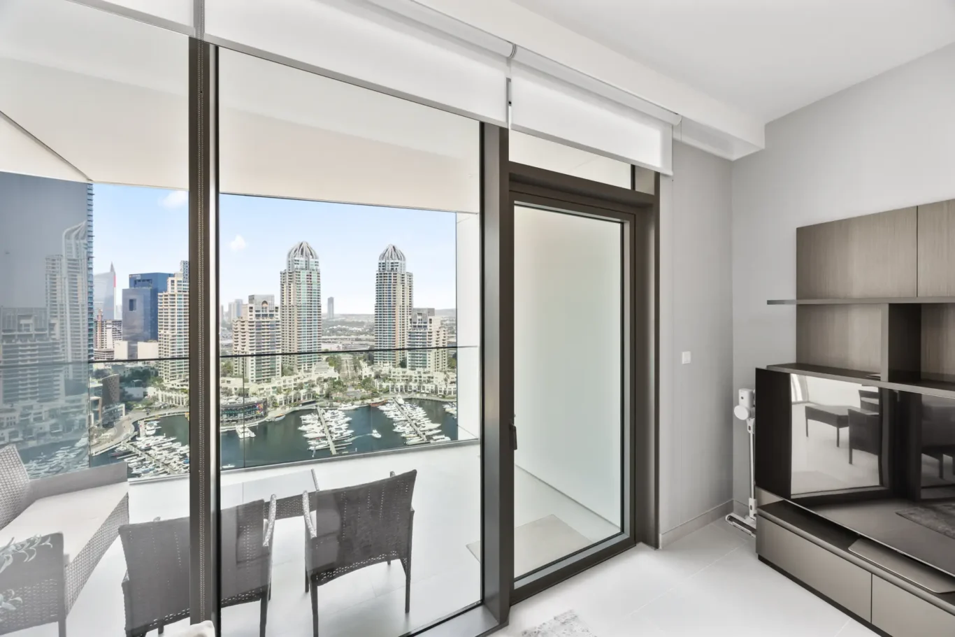 Modern living area with large sliding glass doors opening to a balcony and a harbor view of boats and city skyline behind light-filtering blinds, gray walls, and contemporary cabinetry.