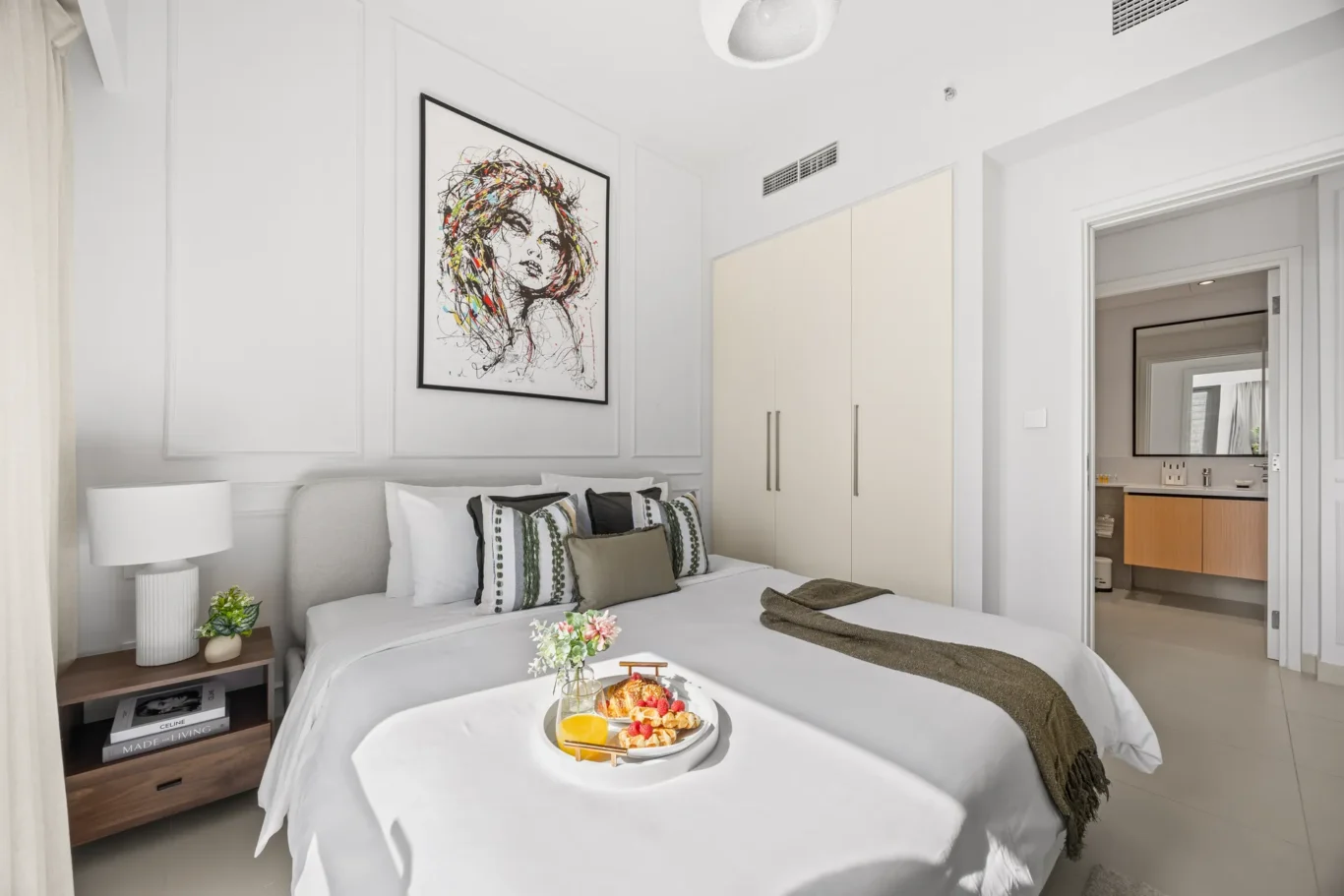 Bright hotel bedroom with white bedding, decorative throw pillows, and a breakfast tray on the bed.