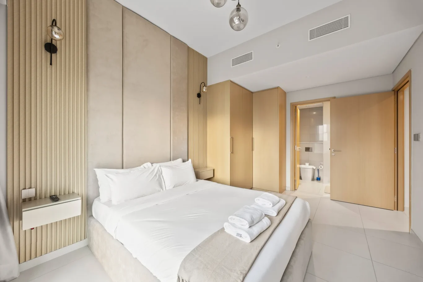Hotel bedroom with a large white bed, beige headboard, and wooden wardrobes nearby the open bathroom door.