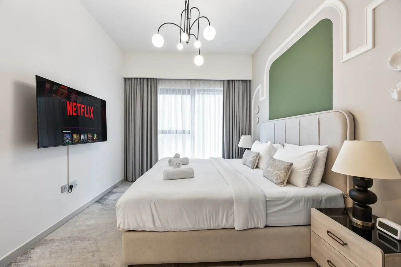 Modern hotel room with a wall-mounted Netflix TV, a large white bed, and a green padded headboard panel.