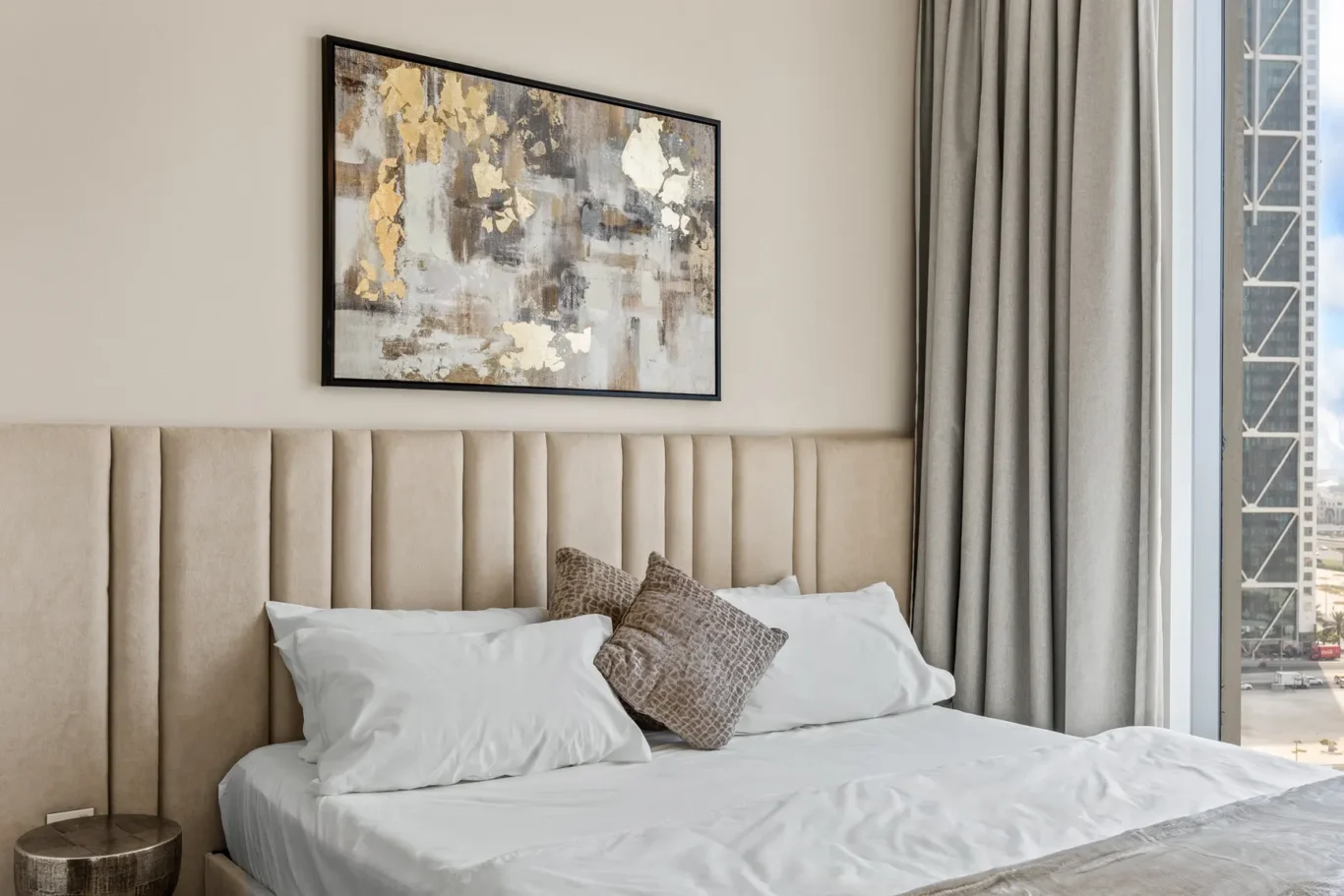 Hotel-style bedroom with a tufted beige headboard, white bedding, and a brown textured throw pillow; abstract art above and gray curtains by a window.