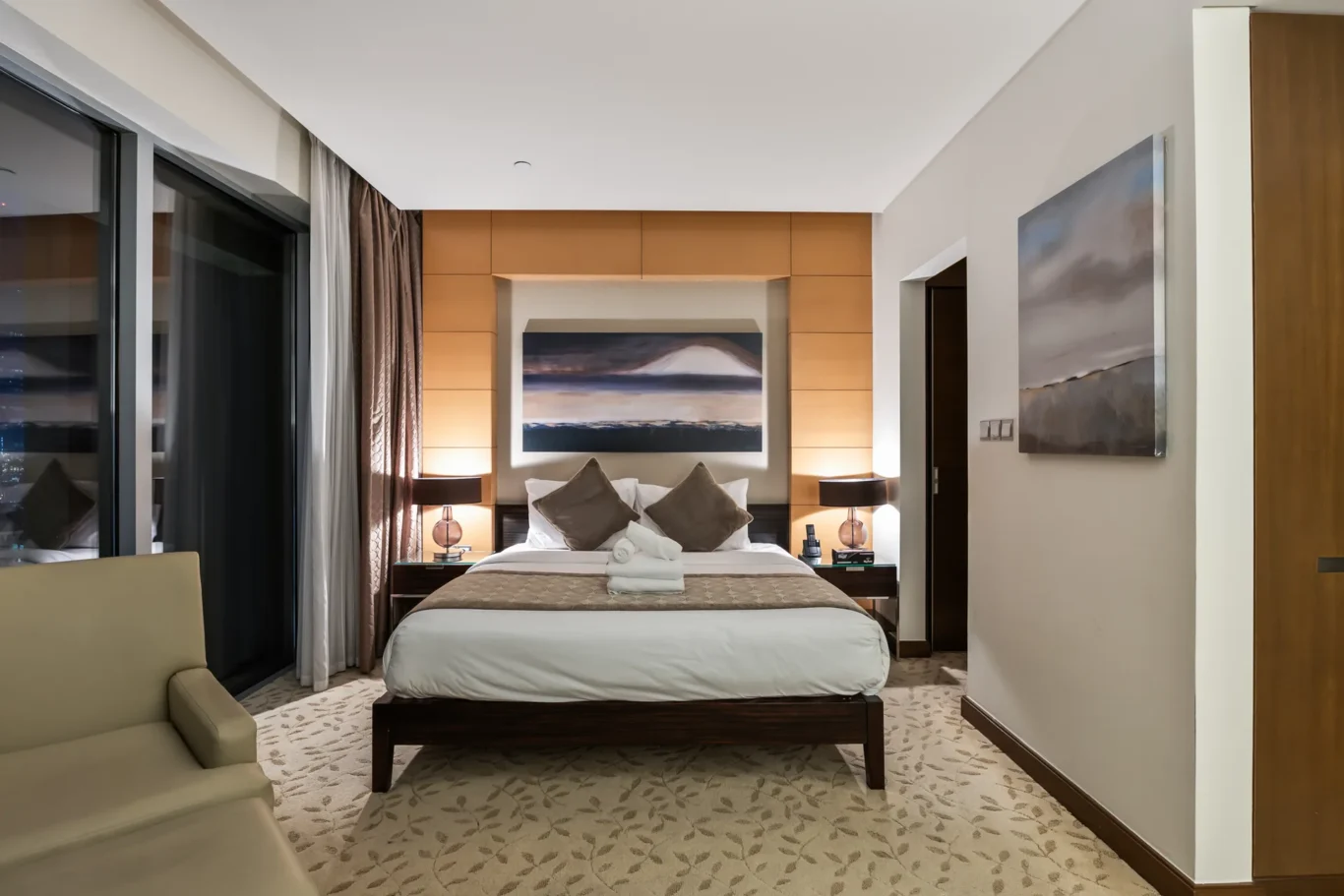 Hotel bedroom with a king bed, neutral bedding, and warm wood headboard panels flanked by two nightstands and lamps.