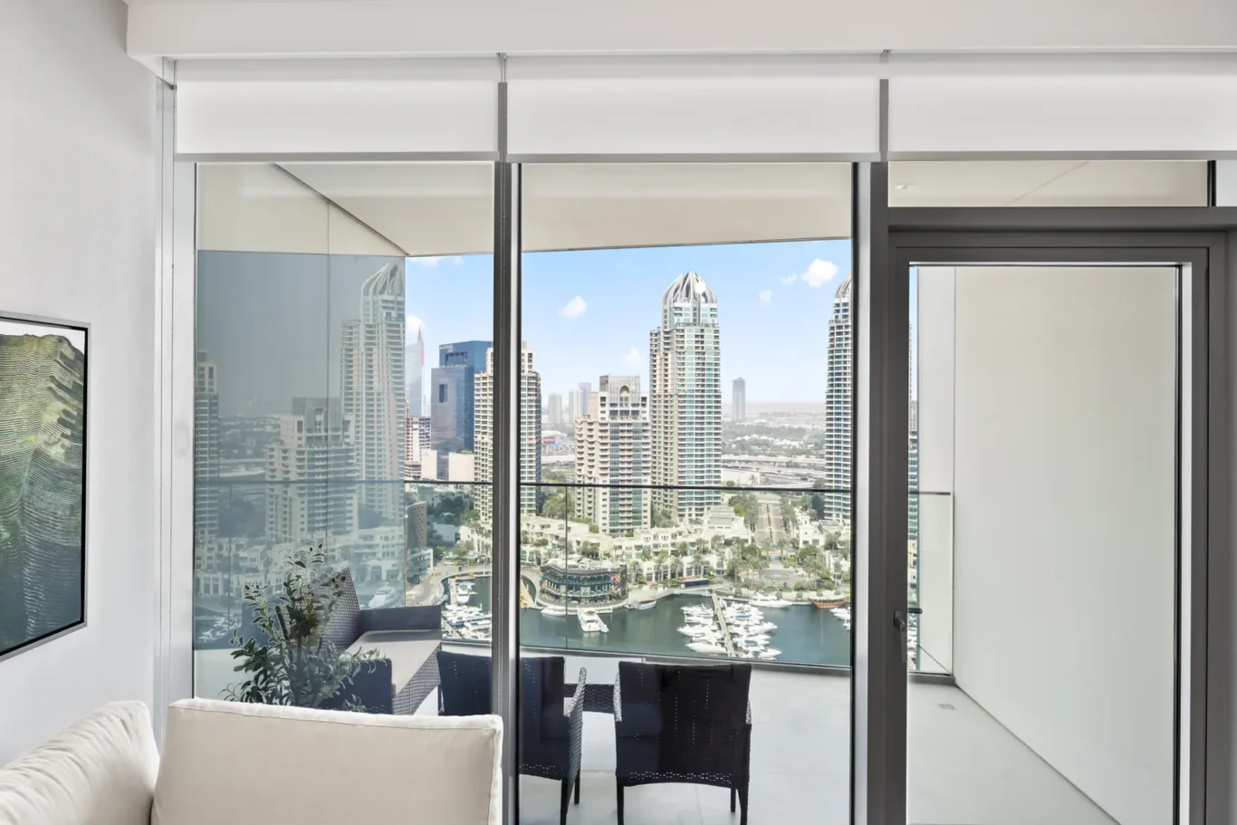 Bright living room with floor-to-ceiling glass doors opening to a balcony, overlooking a marina and city skyline of tall buildings.