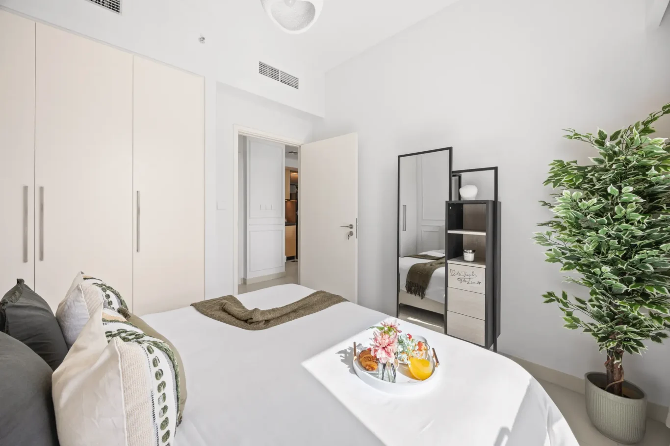 Bright modern bedroom with white wardrobes along left wall, bed in foreground, open door to hallway, and a tall leafy plant on the right.