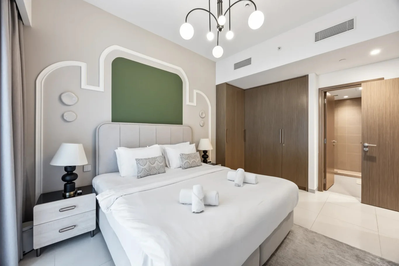 Modern hotel bedroom with two neatly made beds, decorative pillows, and a gray upholstered headboard; green accent wall panel behind the bed and stylish wall molding. A black nightstand and white lamp sit beside each bed, and a doorway to a wood-clad closet and bathroom is visible.