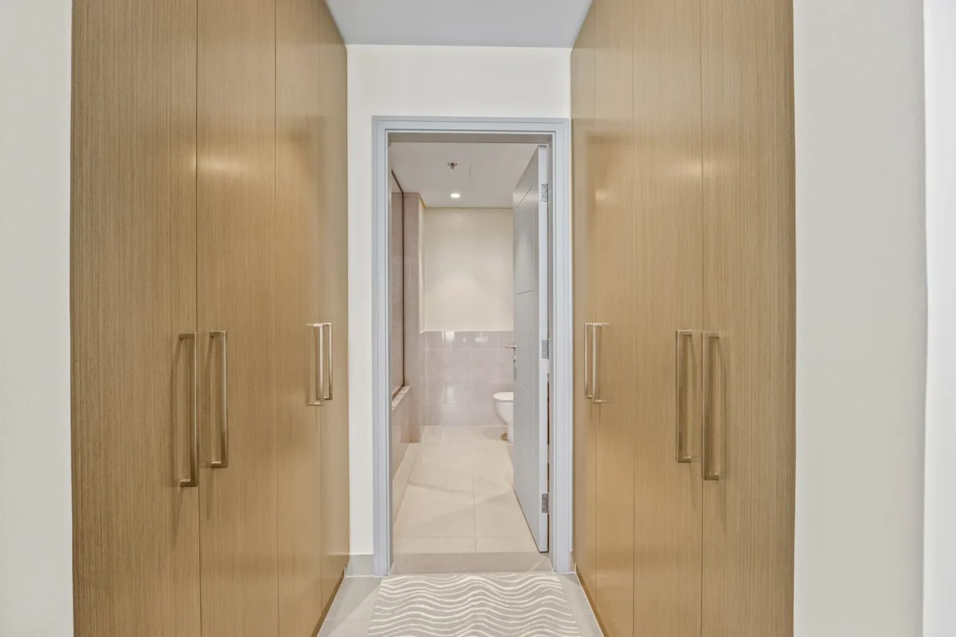 Narrow hallway lined with tall wooden wardrobes on both sides, opening into a light tiled bathroom.