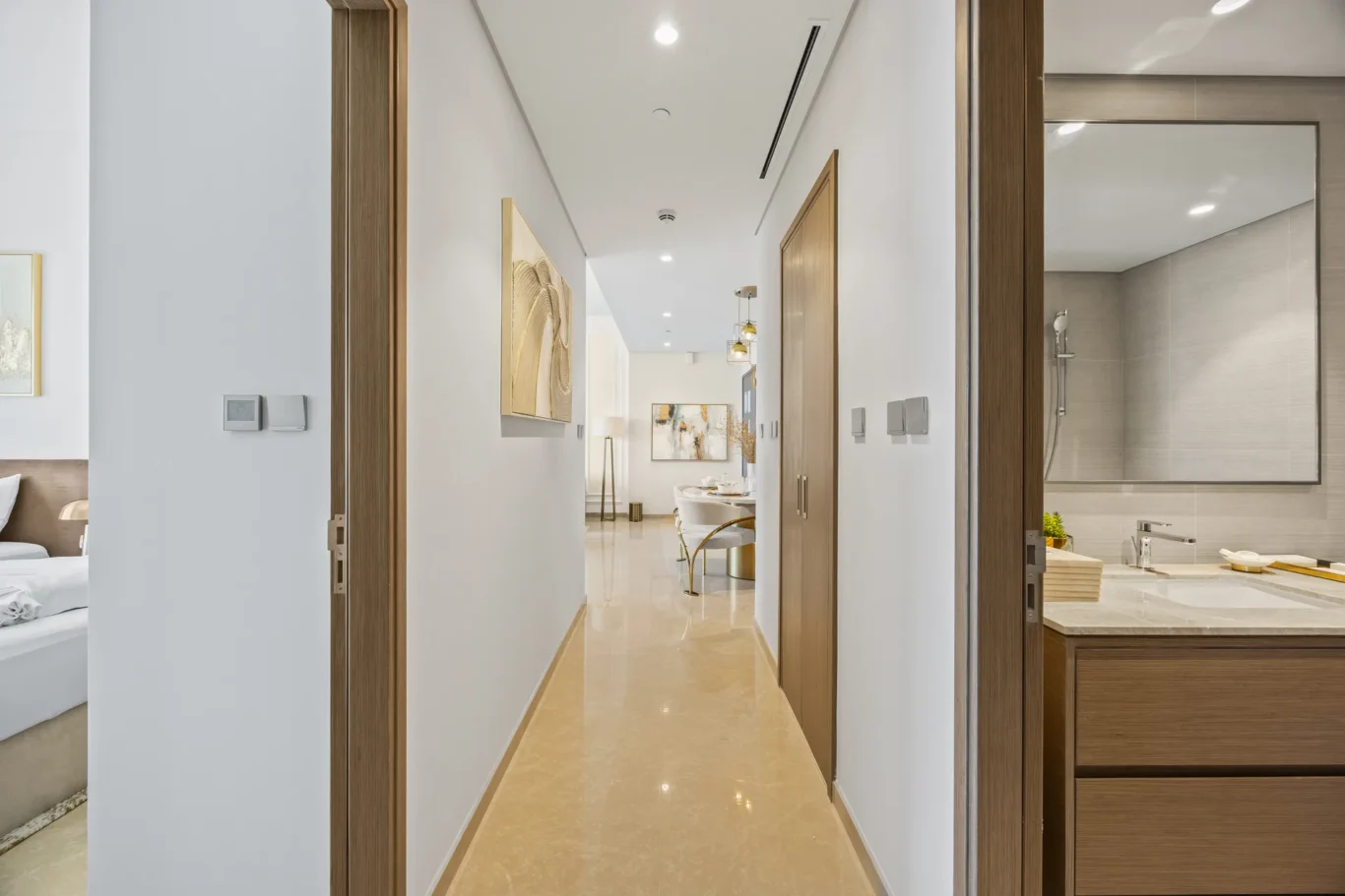 Bright, modern hallway in a home with white walls, wood door frames, and warm lighting; dining area visible straight ahead and a bathroom/kitchen area to the right.