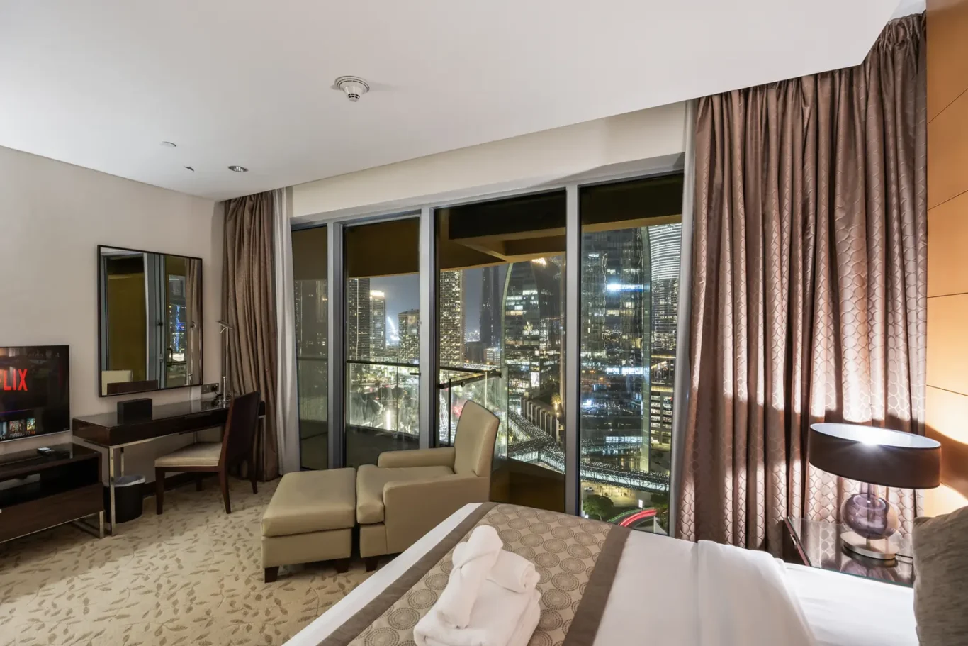Hotel room at night with a king bed, beige seating area, and floor-to-ceiling windows showing a lit city skyline outside the balcony.