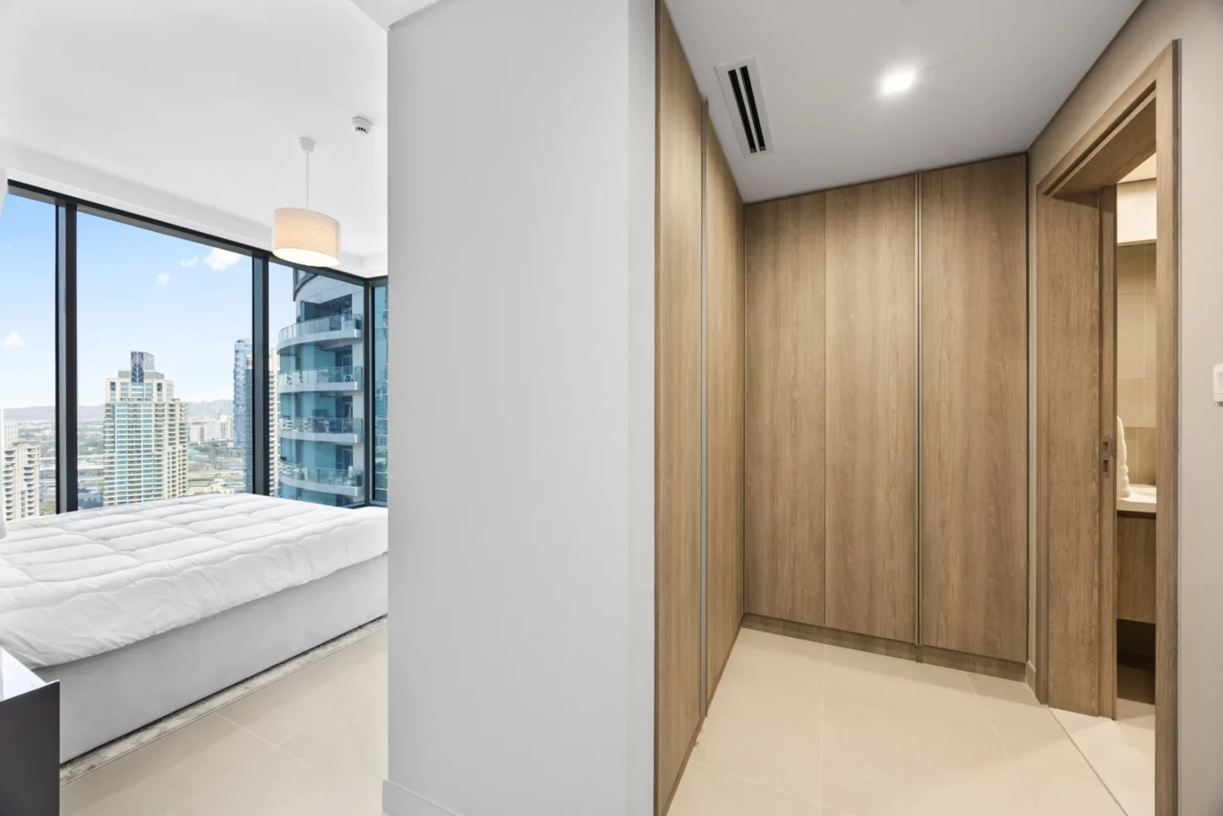 Bedroom with large windows showing a city skyline; white bed on the left and tall wooden wardrobes on the right.