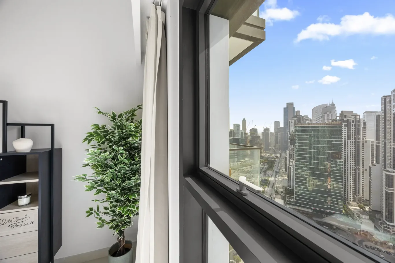 Interior living space with a tall potted plant by a large window and white curtains; city skyscrapers visible outside in daylight.