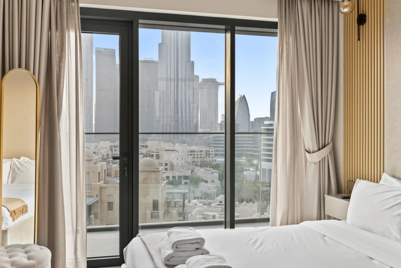 Hotel room with floor-to-ceiling glass balcony doors overlooking a city skyline, white bedding, and folded towels on the bed.