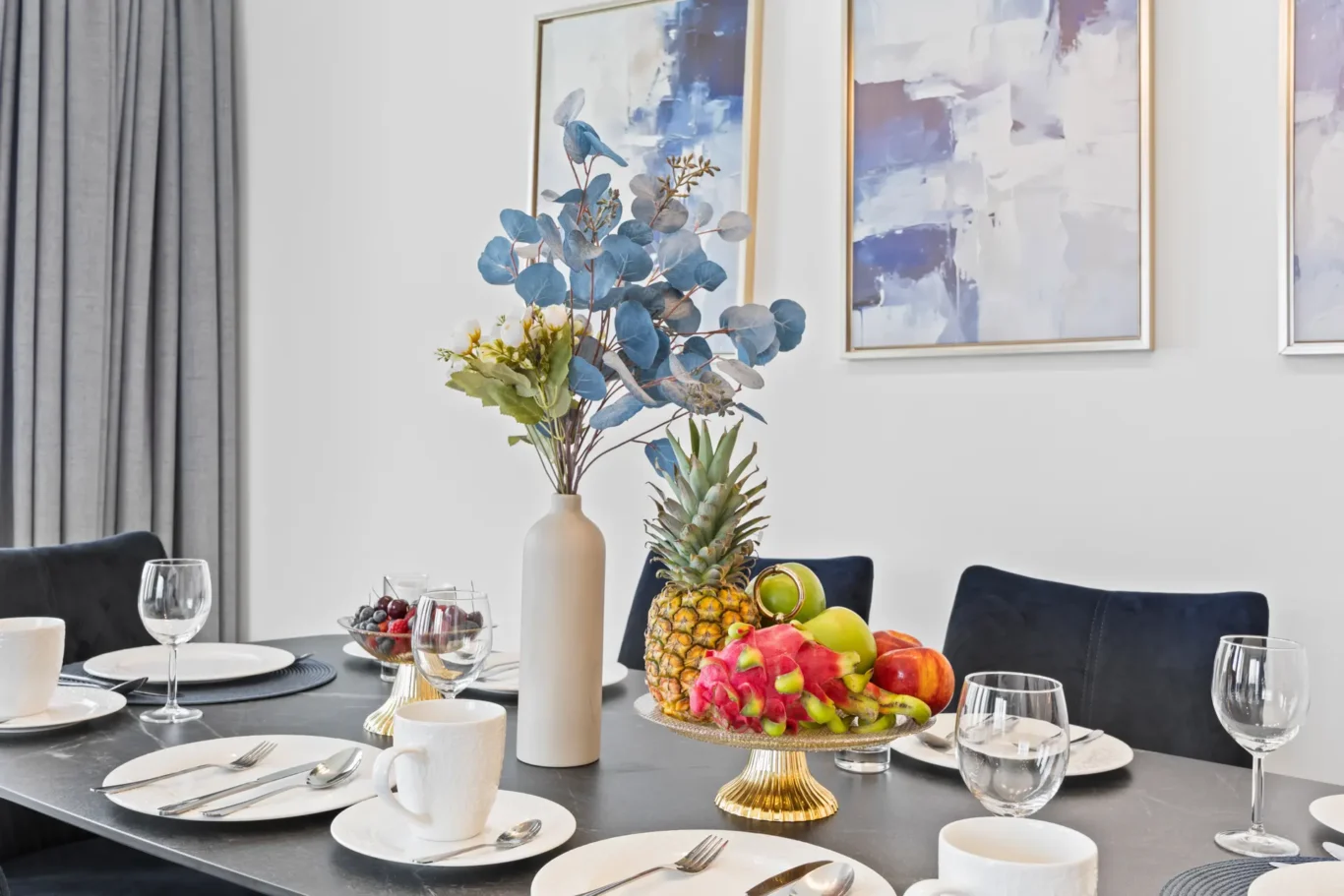 Table set for a meal with plates, mugs, and glasses; centerpiece features a vase of blue eucalyptus beside a fruit platter including pineapple and dragon fruit.