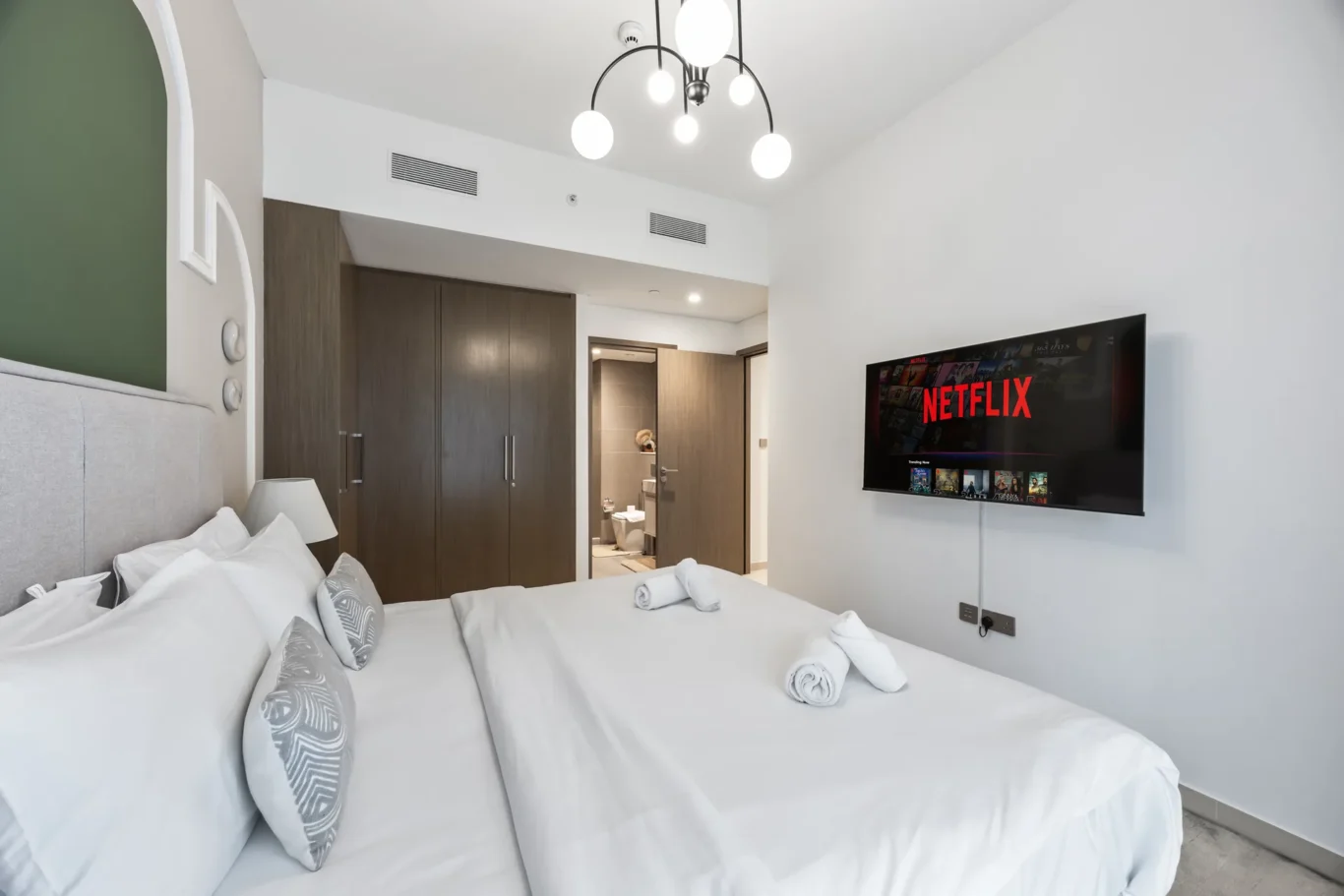 Modern hotel room with a large white bed, pillows and towels, a wall-mounted TV displaying Netflix, and dark wooden wardrobes nearby.