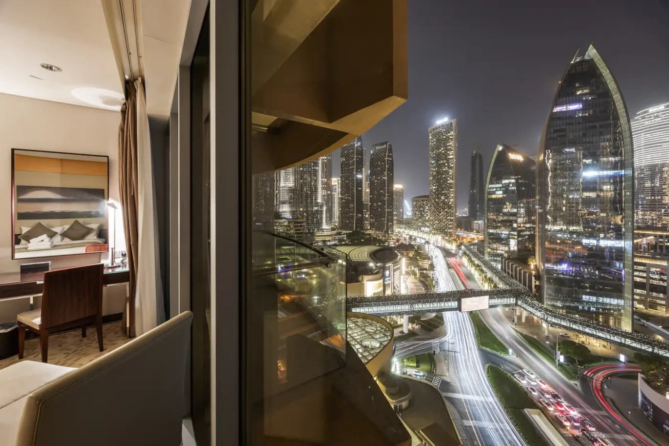 Hotel room interior at night with a desk and chair, and a large window showing a brightly lit city skyline with tall buildings and highway lights outside.