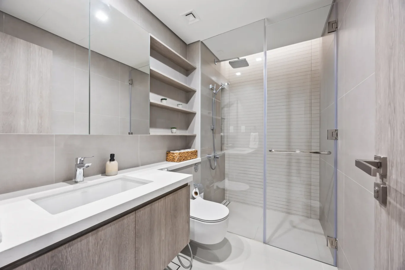 Modern grey-tiled bathroom with a glass-enclosed shower, floating wood vanity, and open shelving.
