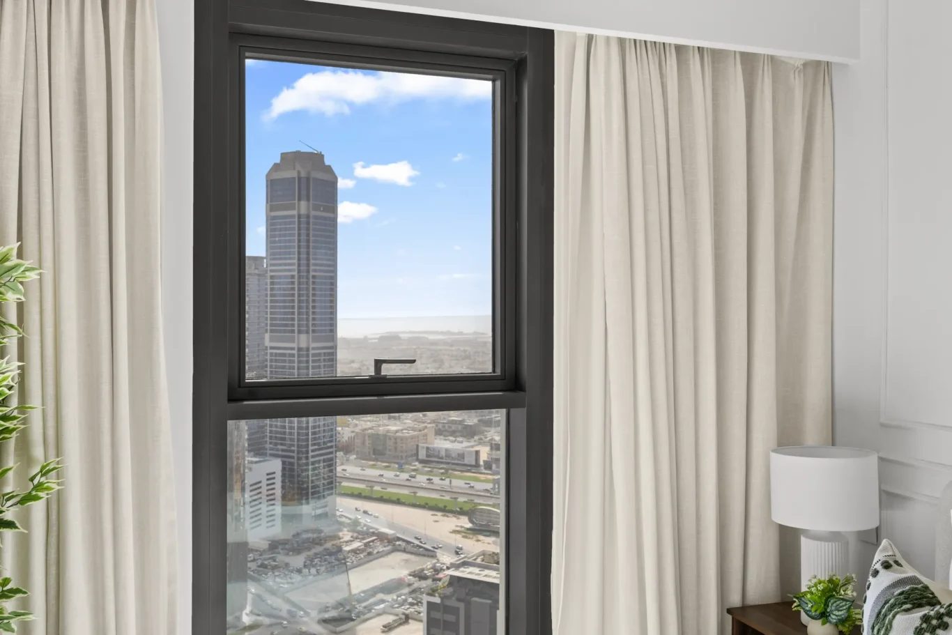Window with black frame flanked by cream curtains; city skyline and blue sky visible outside.