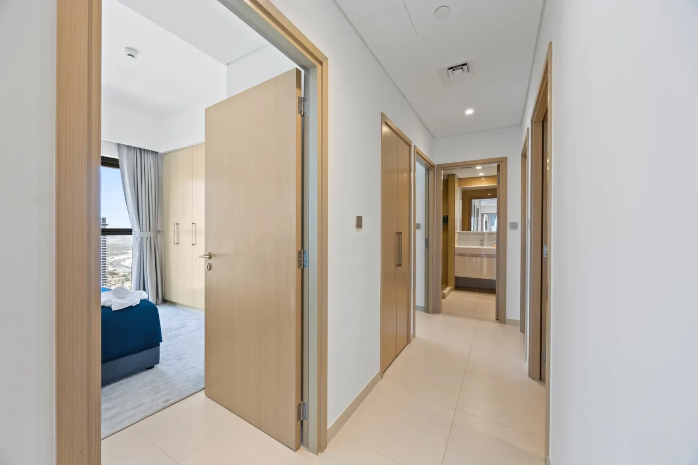 Bright hotel-style hallway with light wood doors and a view into a bedroom showing a bed and window with city scenery outside
