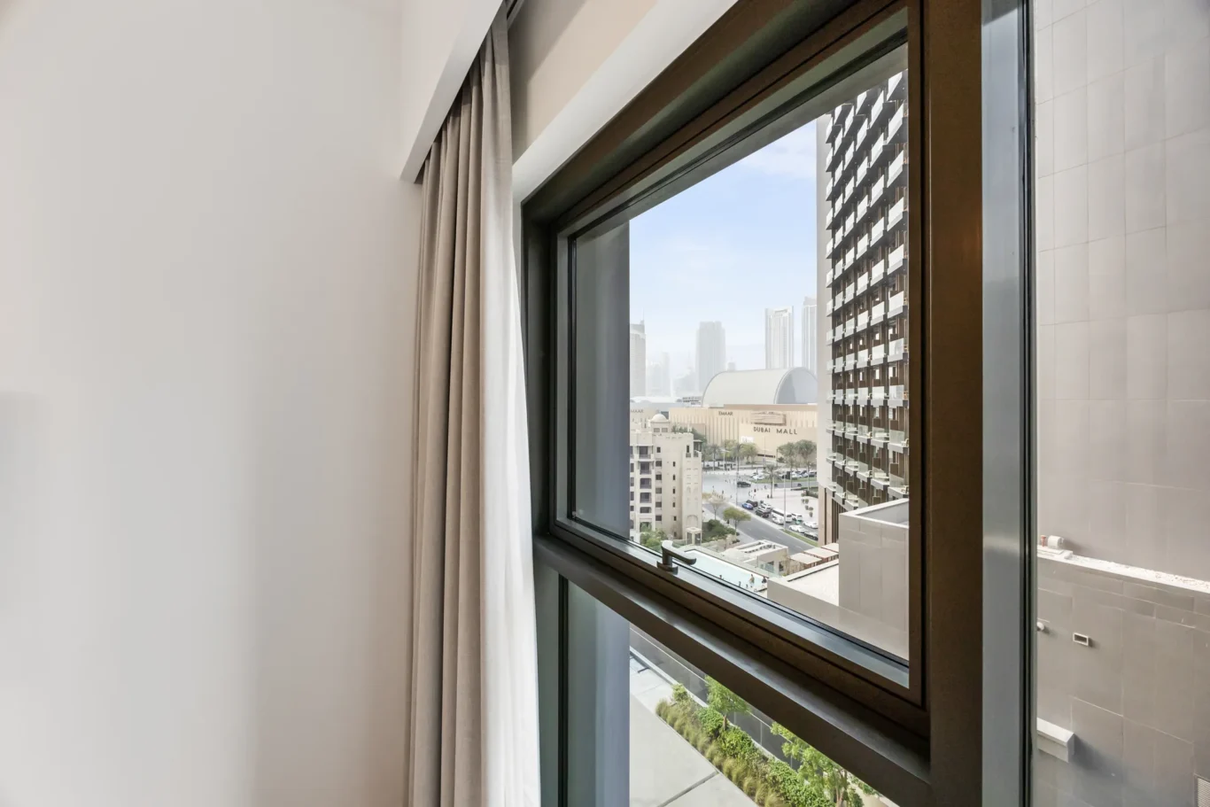 Window with beige curtains, looking out onto a cityscape of high-rise buildings and a mall below.