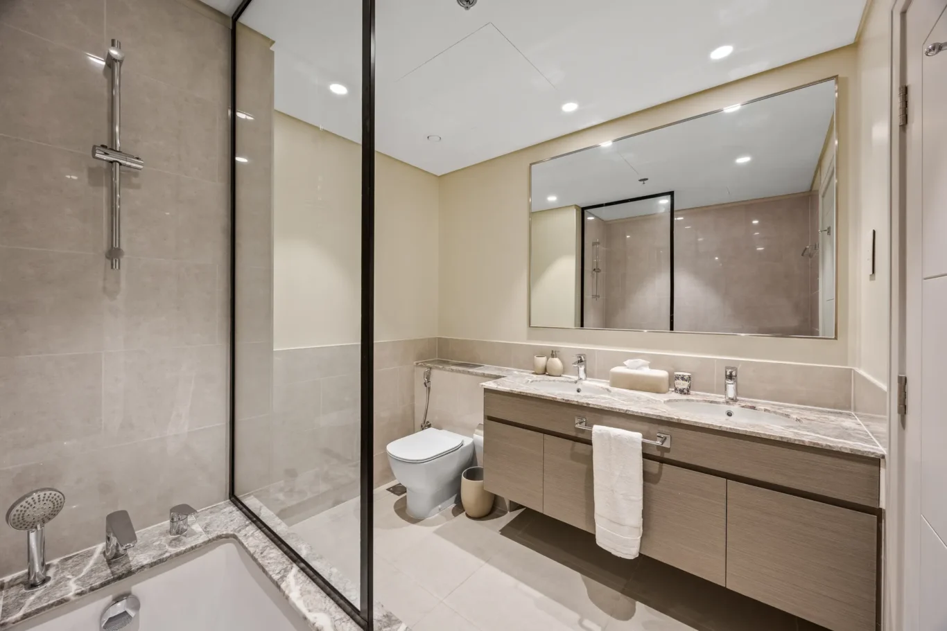 Modern bathroom with a glass-walled shower, a double-sink vanity, and beige tiled walls and floor under bright ceiling lights.