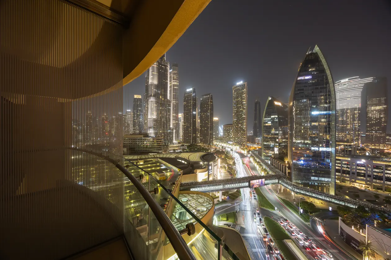 Nighttime city skyline with illuminated skyscrapers and a glass-walled walkway in the foreground.