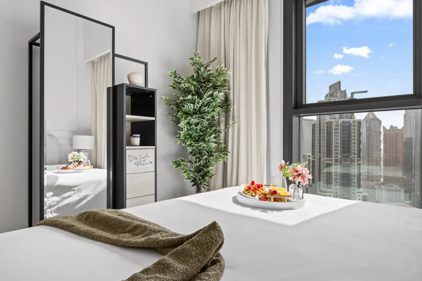 Breakfast tray with waffles and fruit on a neatly made bed, city skyline visible through a large window