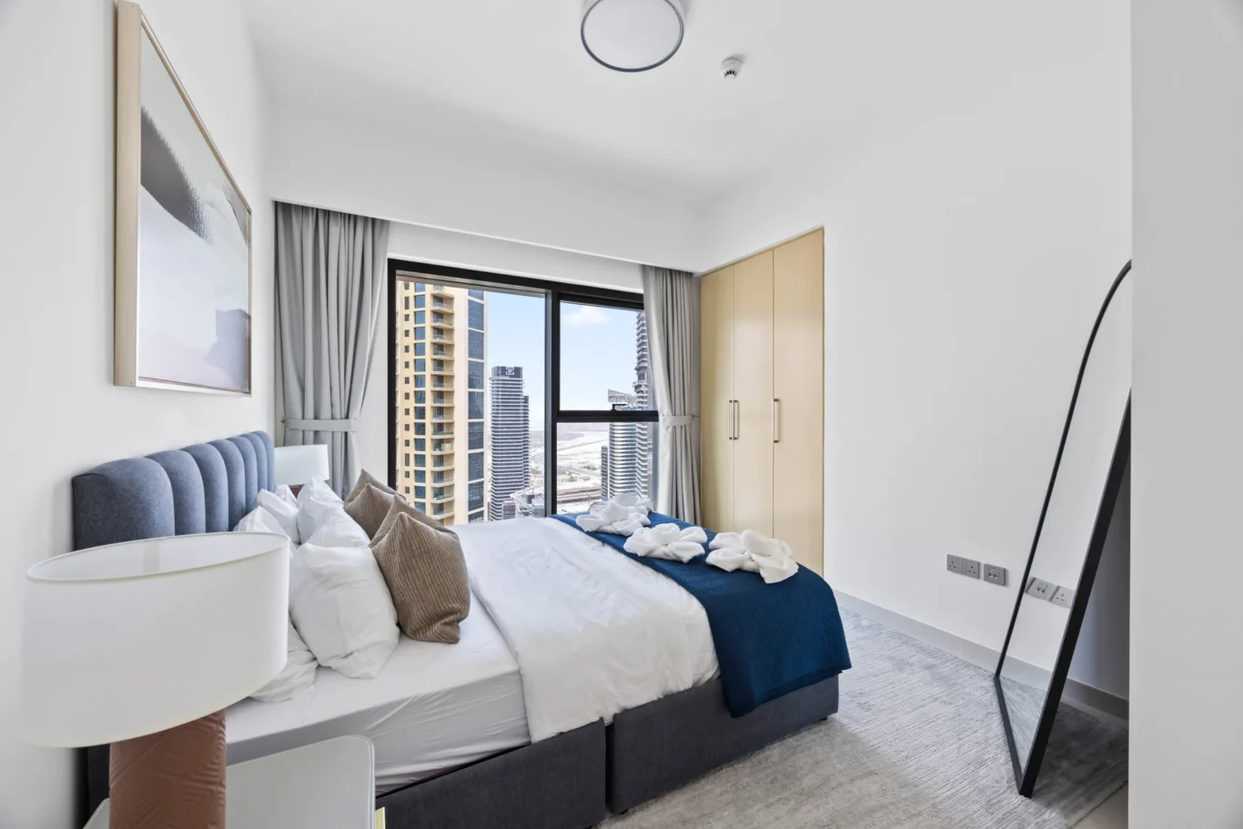 Modern hotel bedroom with white bedding, a blue upholstered headboard, and a city view through a large window.