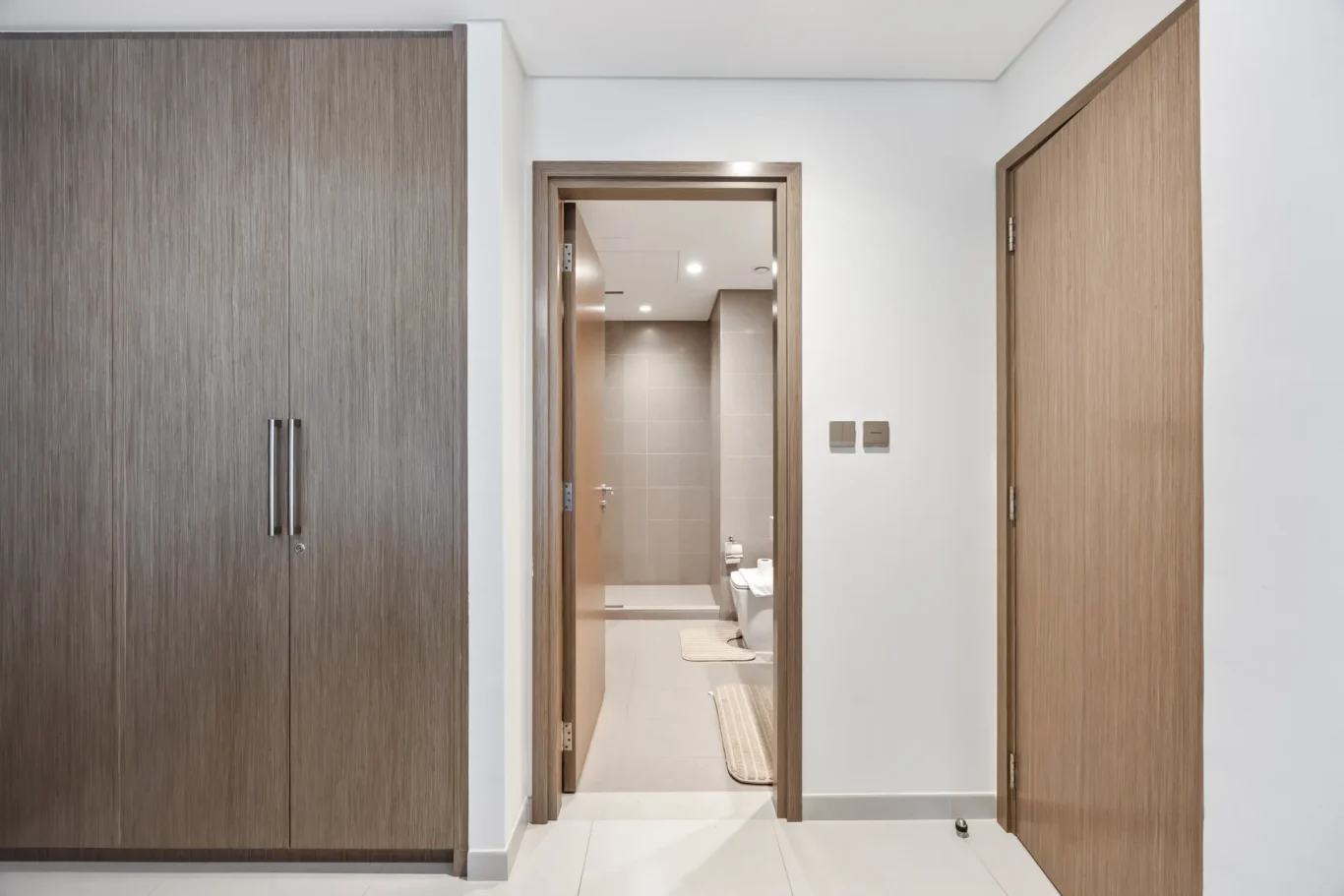 Wide hallway leading to a modern bathroom with gray tiled walls, toilet and bidet visible, flanked by wood-toned doors and a large wardrobe on the left.