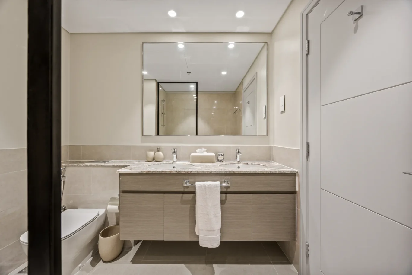 Modern bathroom with a double-sink vanity, large framed mirror, and beige tiled walls.