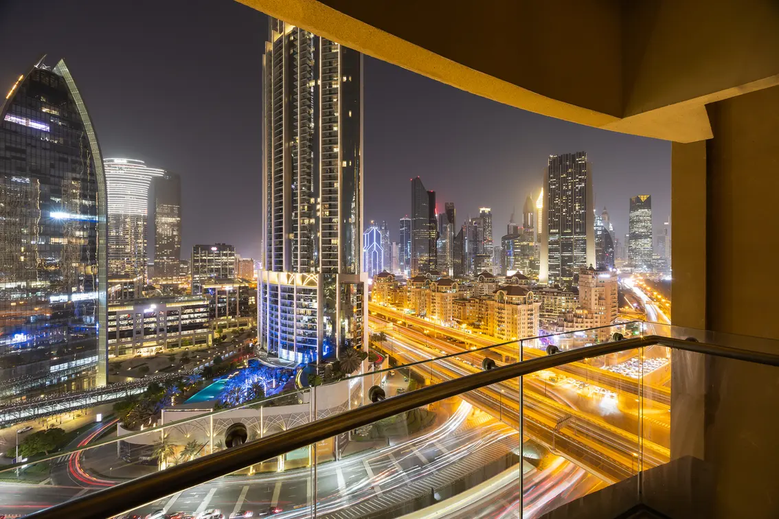 Nighttime city skyline view from a balcony, with illuminated skyscrapers and glowing highways below.