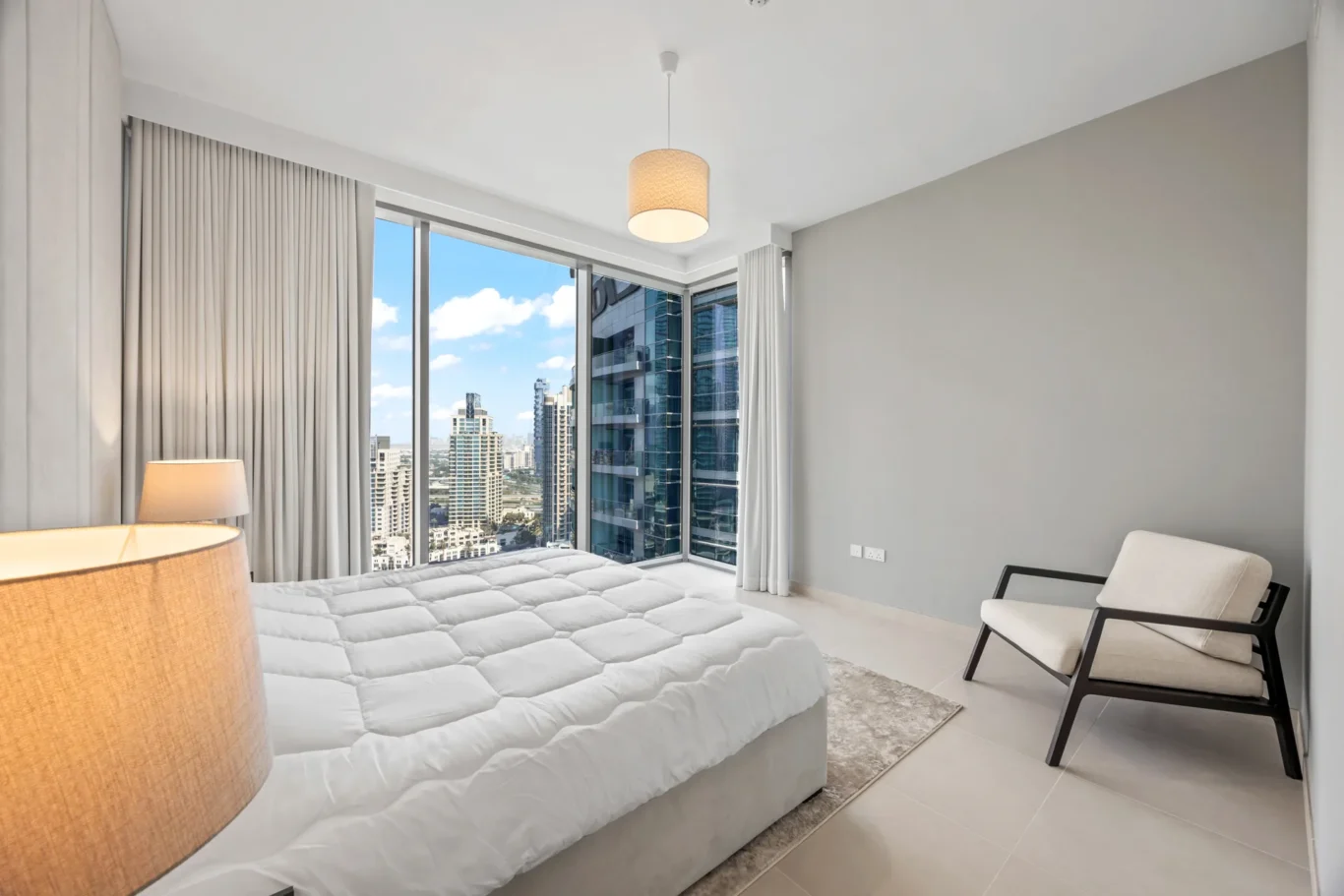 Bright, modern bedroom with a large window showing a city skyline, white bedding, beige lamp, and neutral decor.