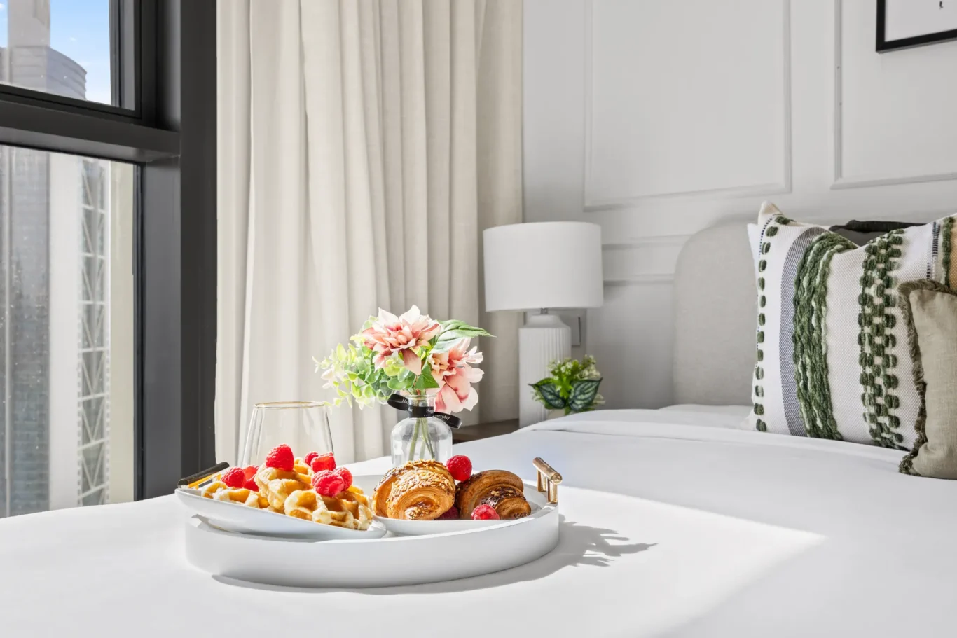 Breakfast spread on a white table in a bright hotel room, with pastries, berries, and a vase of pink flowers near a window.