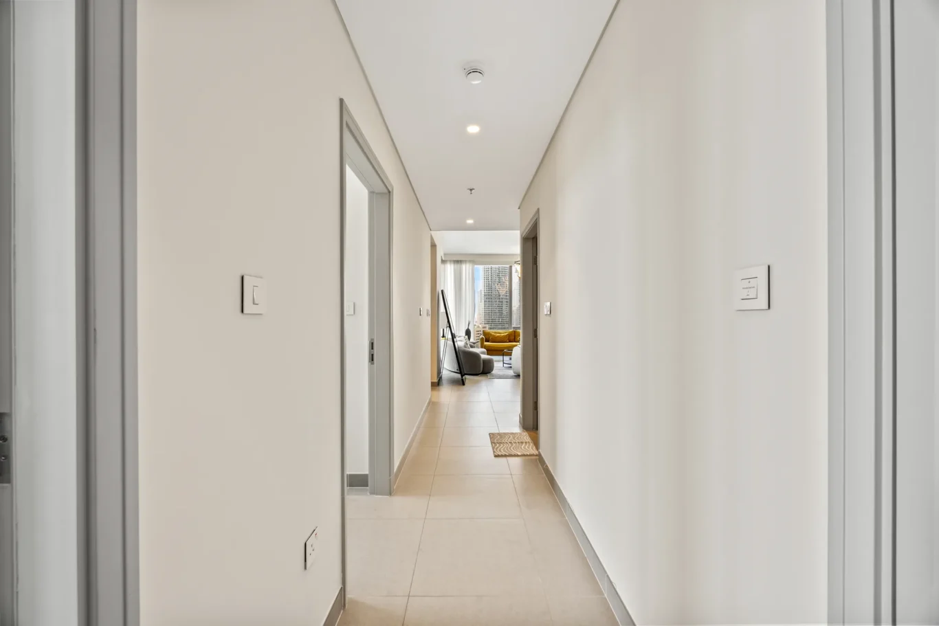 Neutral hallway in a modern apartment with tiled floor, leading to a living area with a mustard sofa at the end.