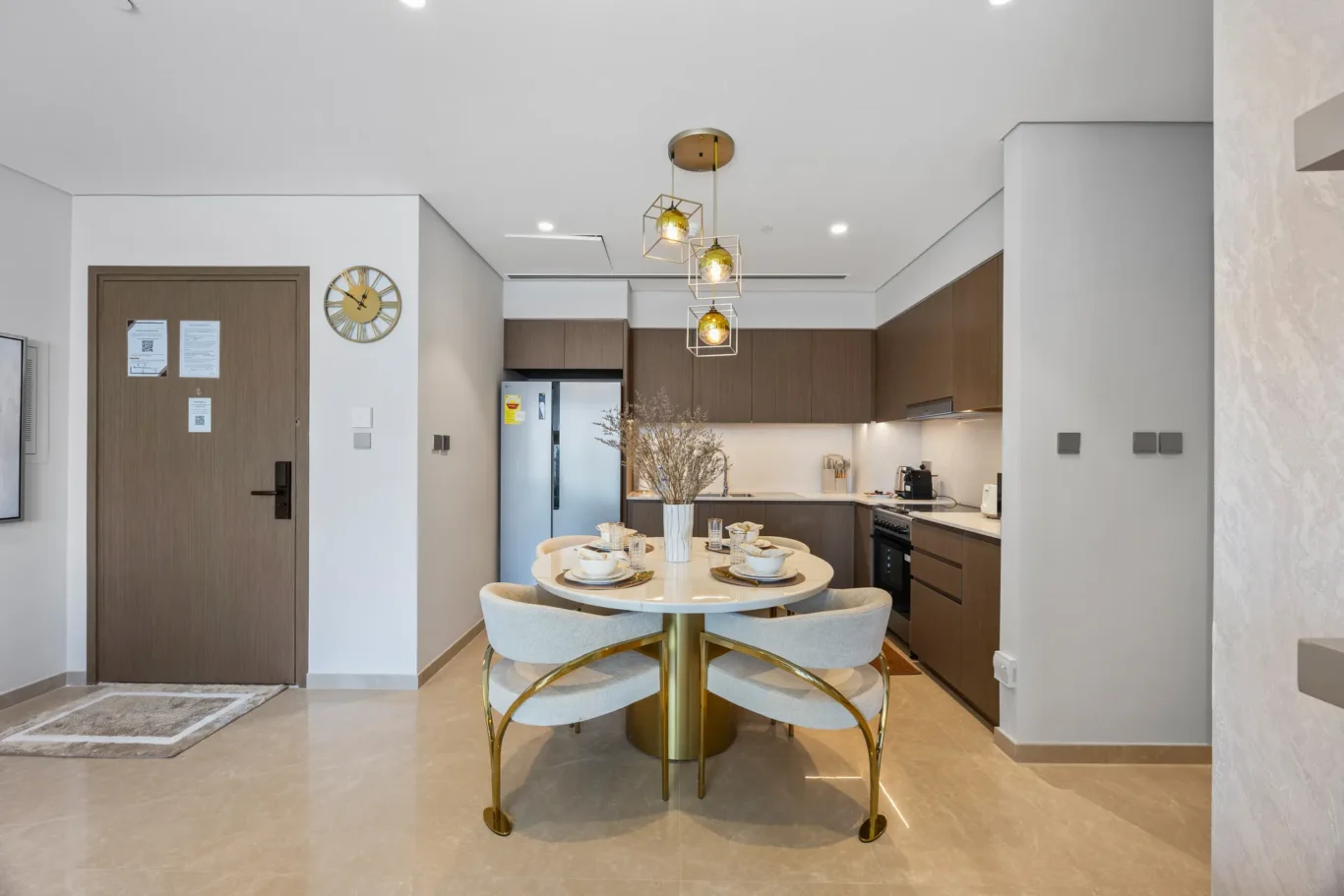 Open-plan kitchen and dining area with a round table set for four, gold-legged chairs, and geometric pendant lights over the table.