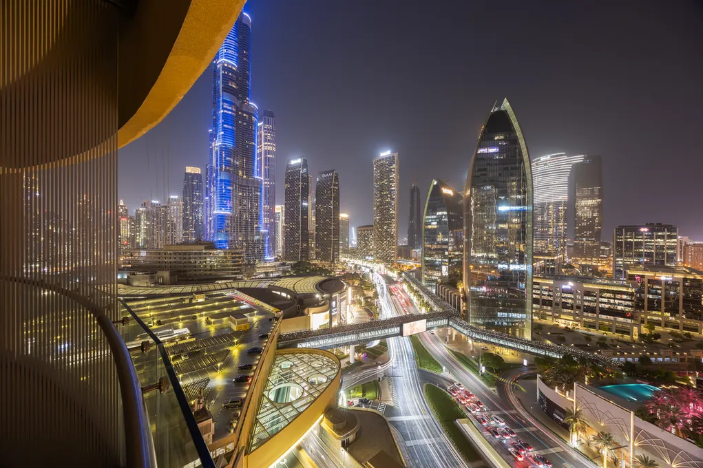 Night view of Dubai skyline at night with illuminated skyscrapers, a curved pedestrian bridge, and traffic on a glowing road below.