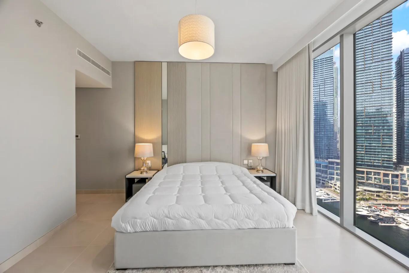 Hotel bedroom with a gray upholstered headboard, white quilted bed, two bedside lamps, and floor-to-ceiling windows offering a city skyline view with tall glass buildings.