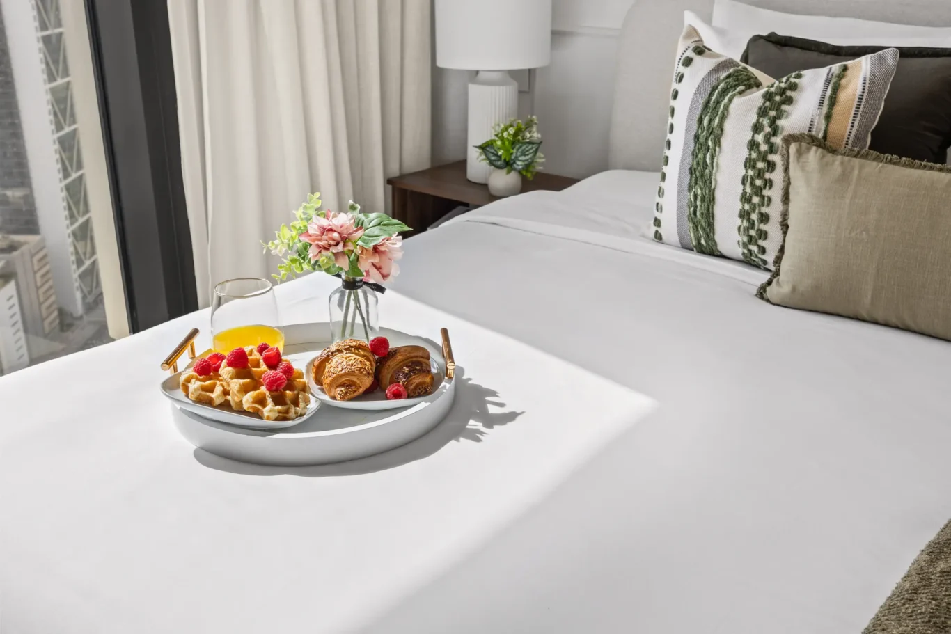 Breakfast on a hotel bed: two plates with waffles, croissants, raspberries, and orange juice beside a small vase of flowers on a white tray by a sunlit window.