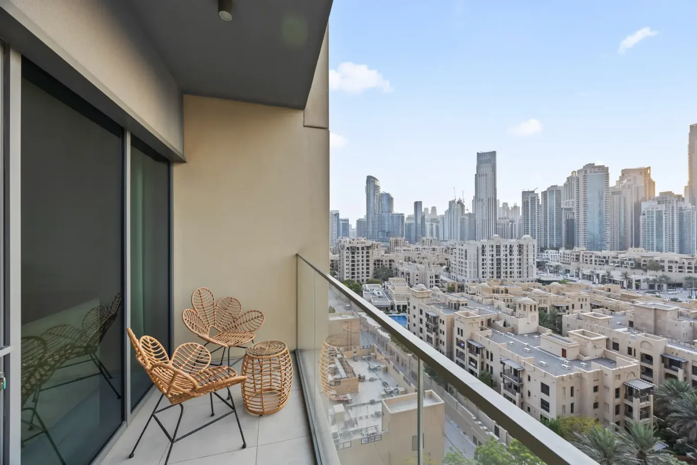 Balcony seating area with woven rattan chairs and a small table, glass railing overlooking a city of high-rise buildings on a sunny day.