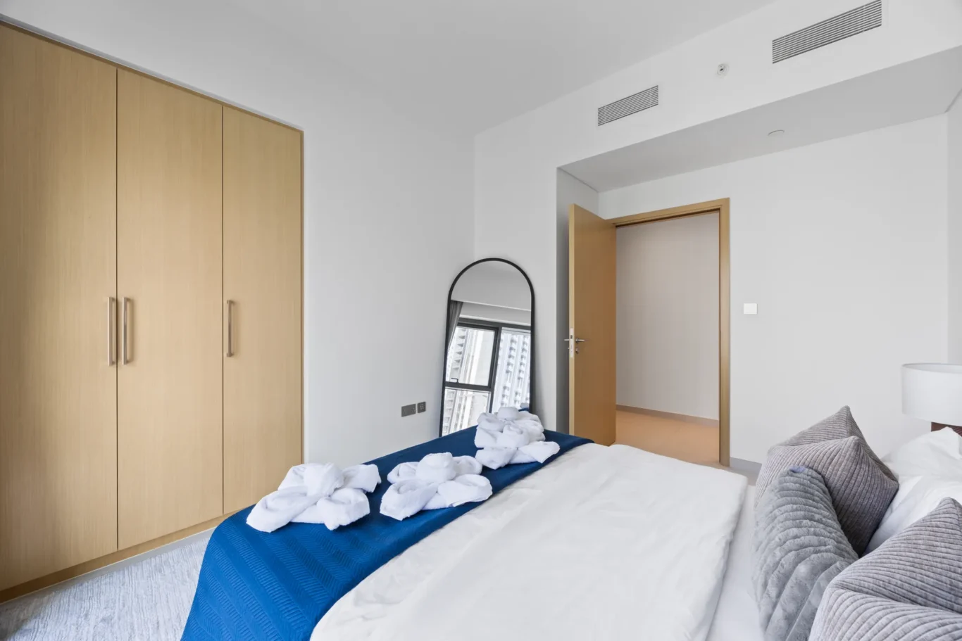 Modern bedroom with light wood wardrobes on the left, white walls, and a bed with a blue runner and folded towels on top.
