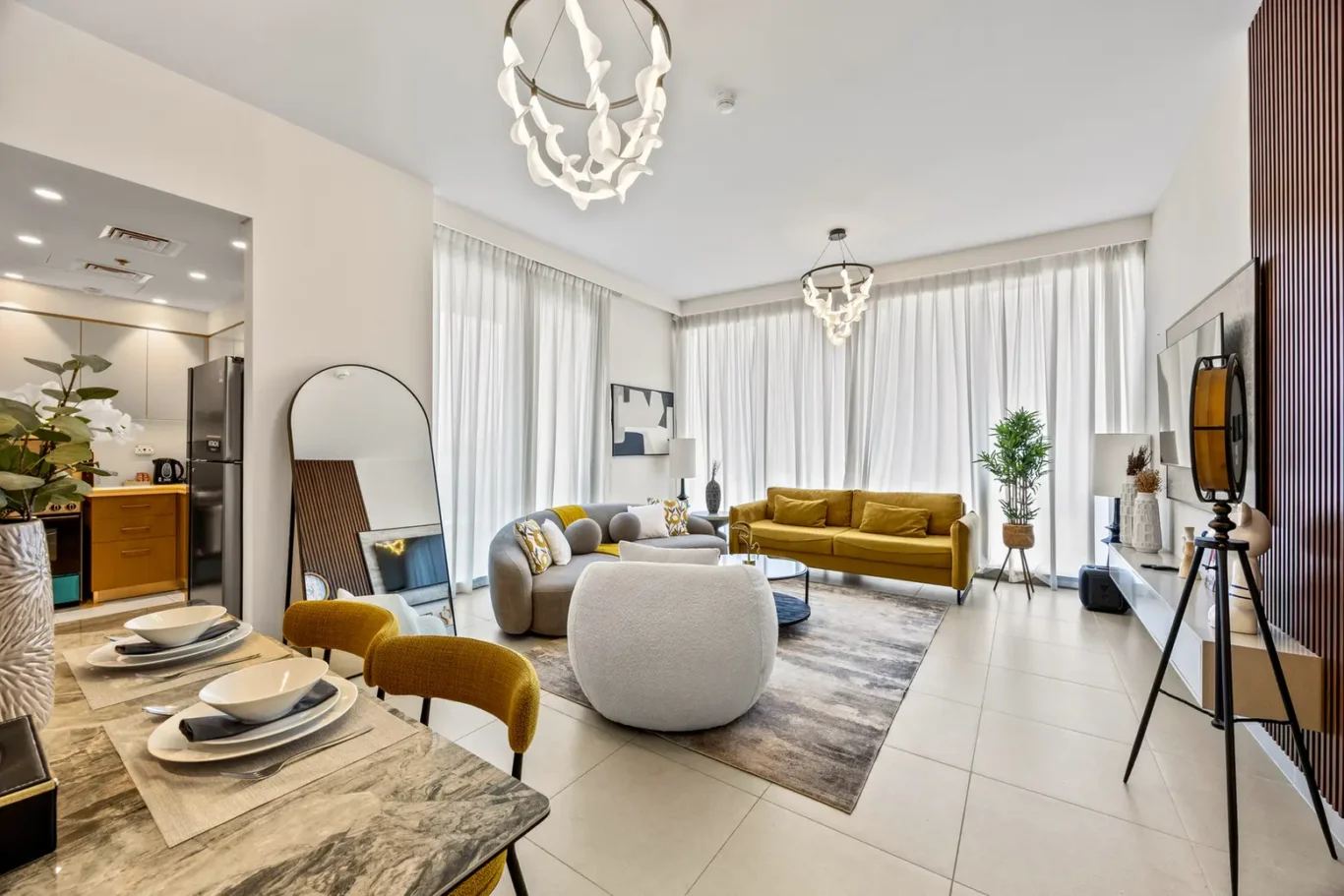 Open-plan living area with a mustard-yellow sofa, gray seating, and a marble dining table near large windows with sheer curtains.