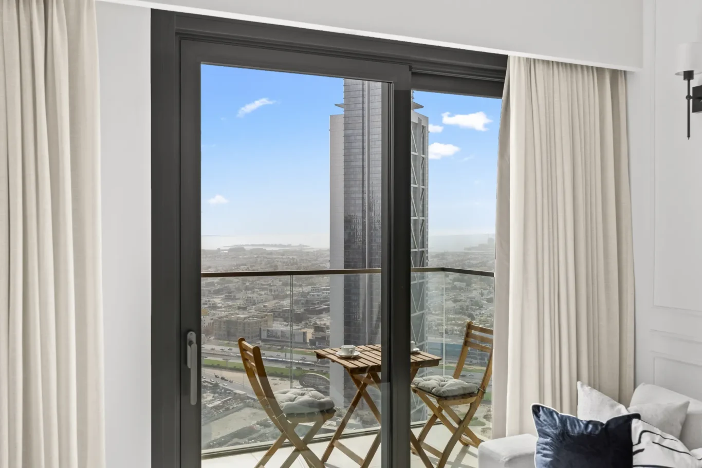Sliding glass door opens to a balcony with a small wooden table and two chairs, overlooking a cityscape and tall buildings.