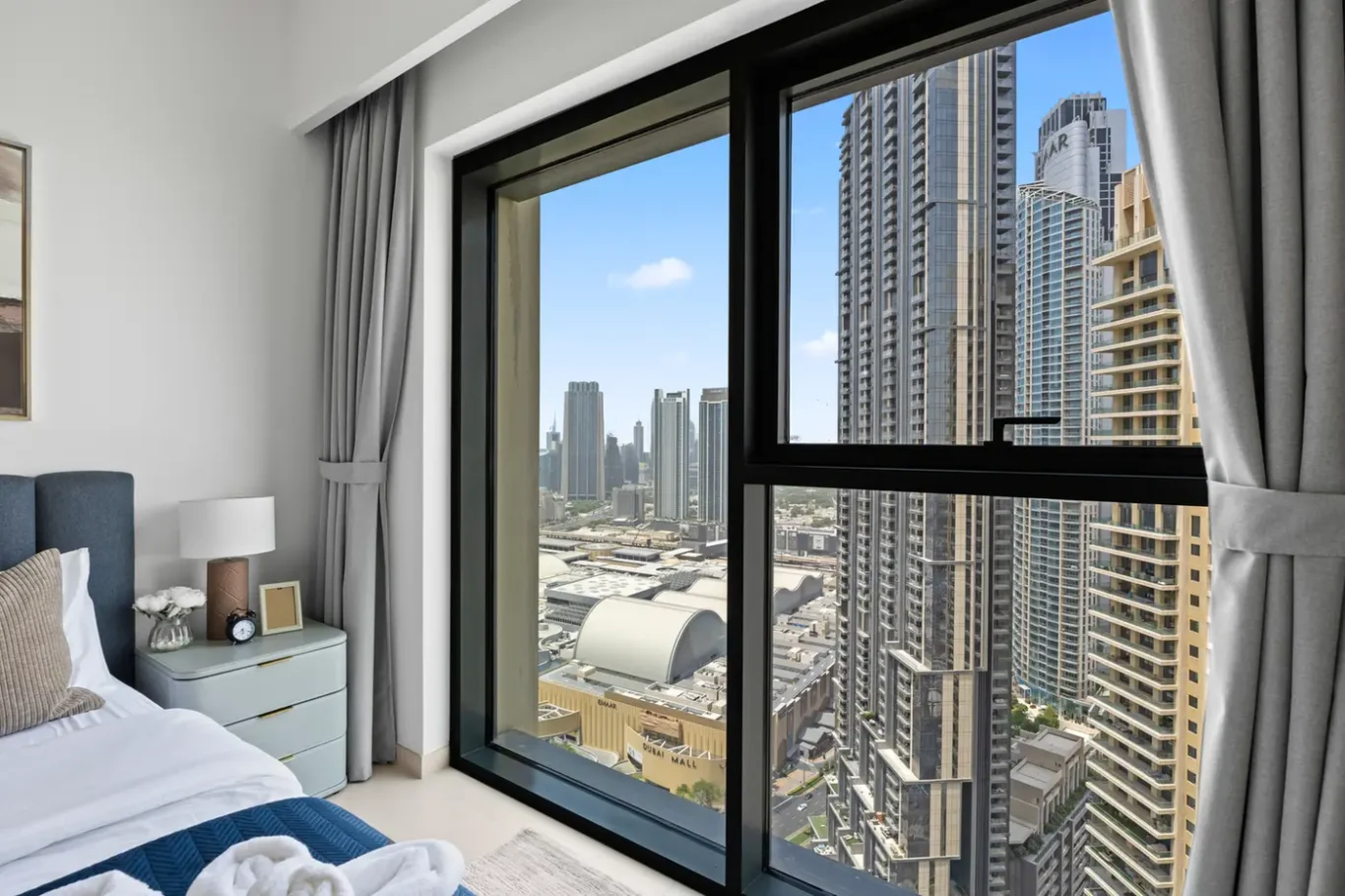 Bedroom with a large window showing a city skyline of tall modern buildings; blue upholstered headboard and nightstand beside the bed.
