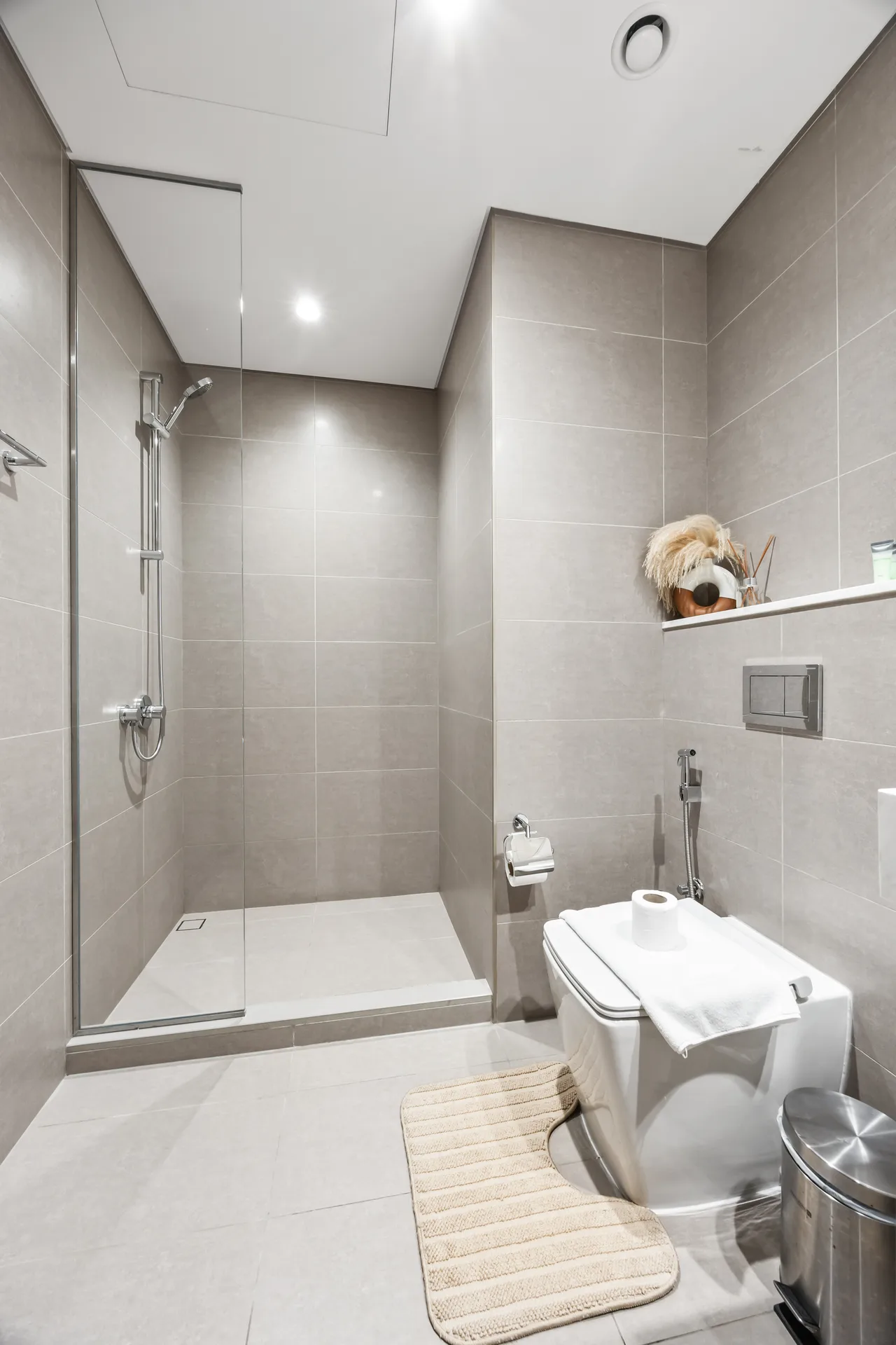 Modern gray-tiled bathroom with a glass shower enclosure on the left and a toilet on the right, beige bath mats on the floor, and a shelf with toiletries.