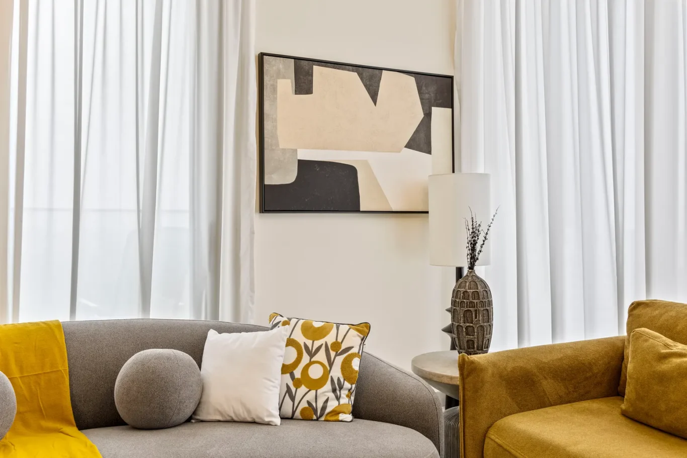Modern living room with a gray curved sofa, mustard throw, and decorative white pillow; abstract wall art above and sheer white curtains.
