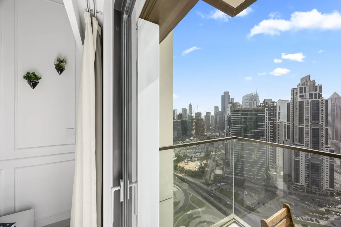 Open sliding glass door leads to a balcony with a glass railing, overlooking a city skyline of tall buildings under a blue sky.