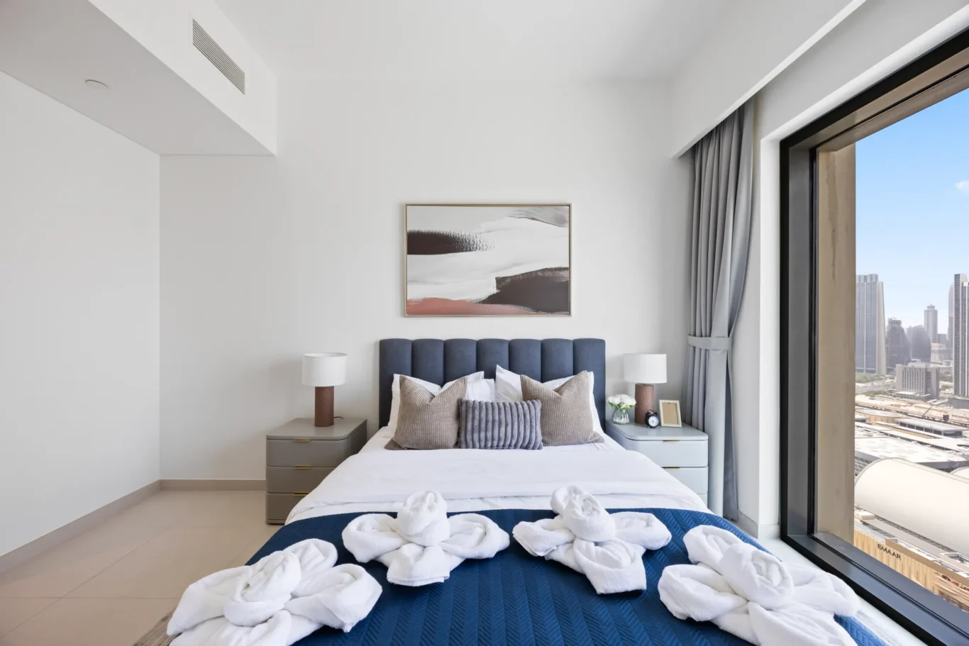 Modern bedroom with blue upholstered headboard, neutral pillows, two gray nightstands, and a large window overlooking the city.